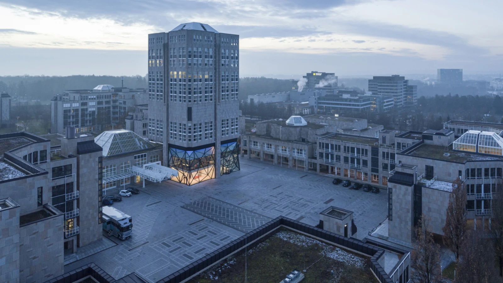Luftaufnahme der Sternhöhe in Stuttgart-Möhringen in der Abenddämmerung, mit Turm, beleuchtetem Glasfoyer und großem Platz